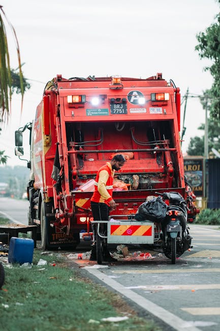 Two mobile waste collection workers are walking across a city street, pushing a wheeled bin filled with discarded materials. One worker is dressed in high-visibility orange trousers and jacket with re