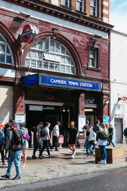 The entrance to Camden Town underground station featuring a red brick façade with arched windows and a prominent blue sign displaying 'Camden Town Station' beneath a roundel London Underground logo. 